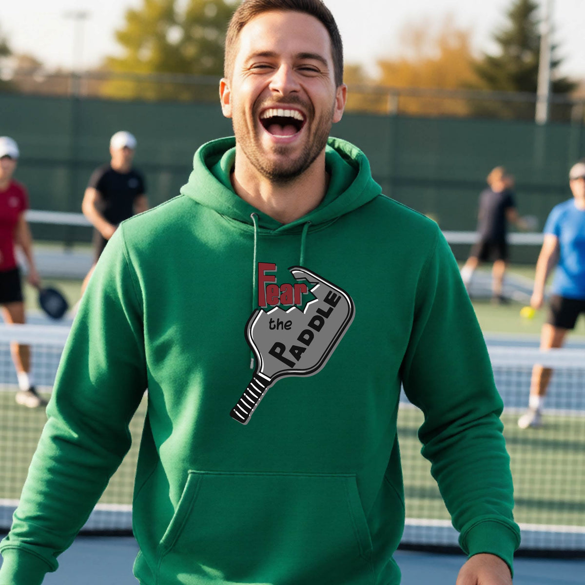 A man wearing a green hoodie pullover featuring a pickleball graphic of the text 'FEAR THE PADDLE' cut into a pickleball paddle, standing on a pickleball court. 