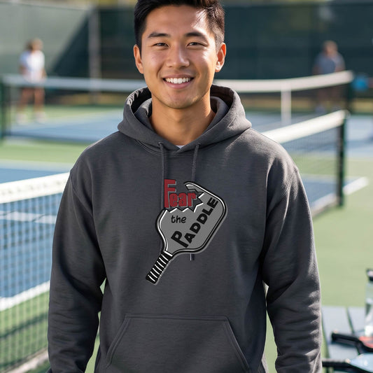 A man wearing a gray hoodie pullover featuring a pickleball graphic of the text 'FEAR THE PADDLE' cut into a pickleball paddle, standing on a pickleball court. 