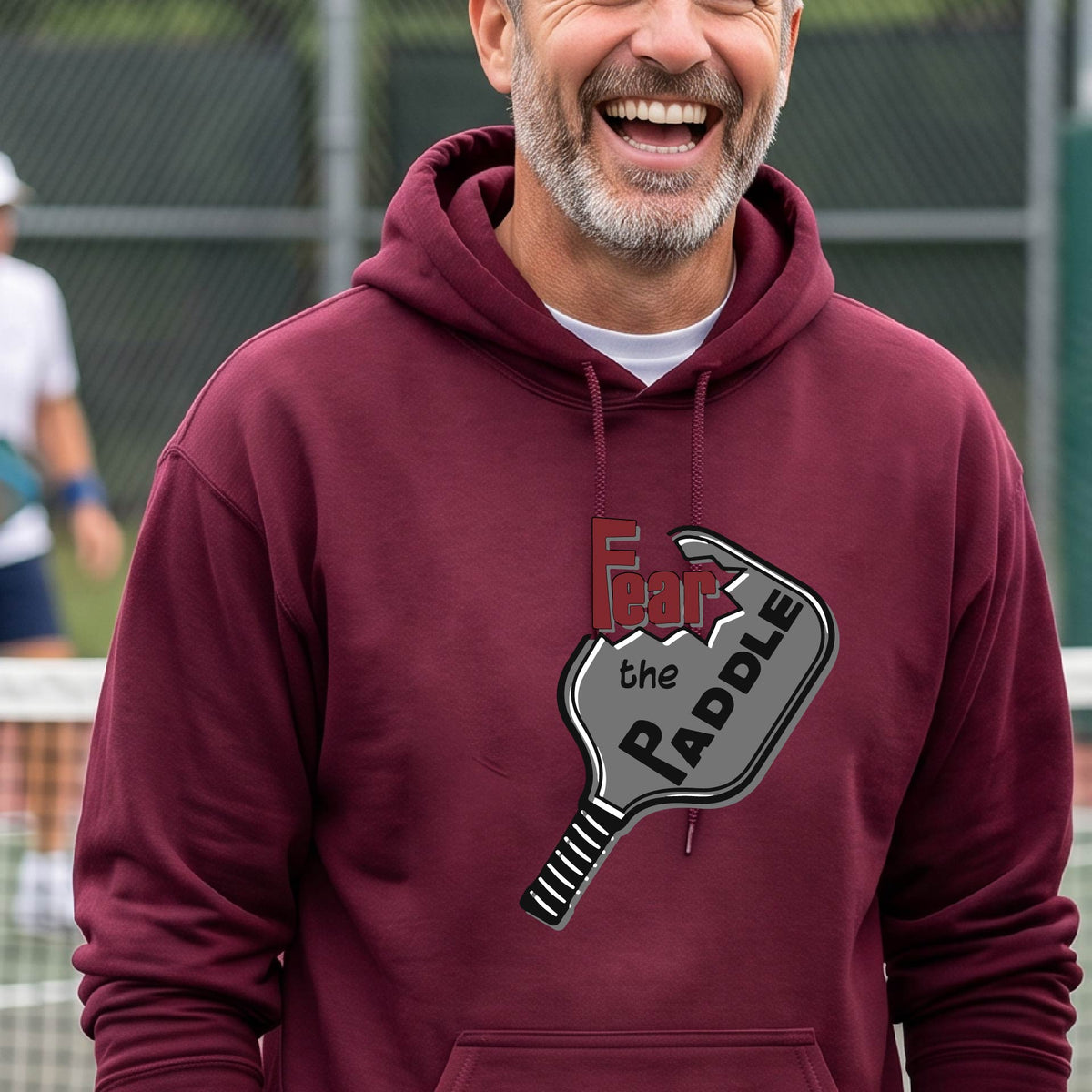 A man wearing a maroon hoodie pullover featuring a pickleball graphic of the text 'FEAR THE PADDLE' cut into a pickleball paddle, standing on a pickleball court. 