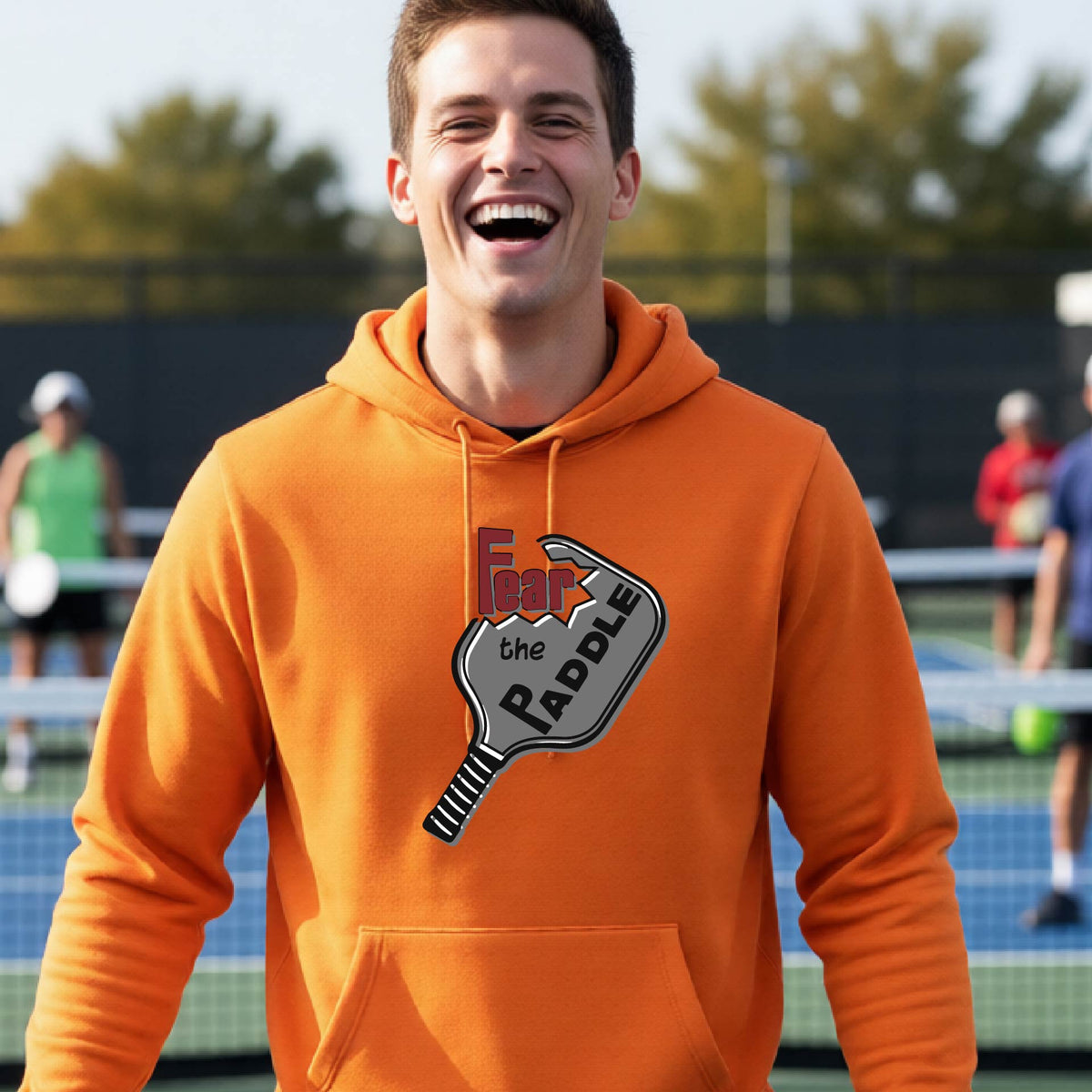 A man wearing a bright orange hoodie pullover featuring a pickleball graphic of the text 'FEAR THE PADDLE' cut into a pickleball paddle, standing on a pickleball court. 