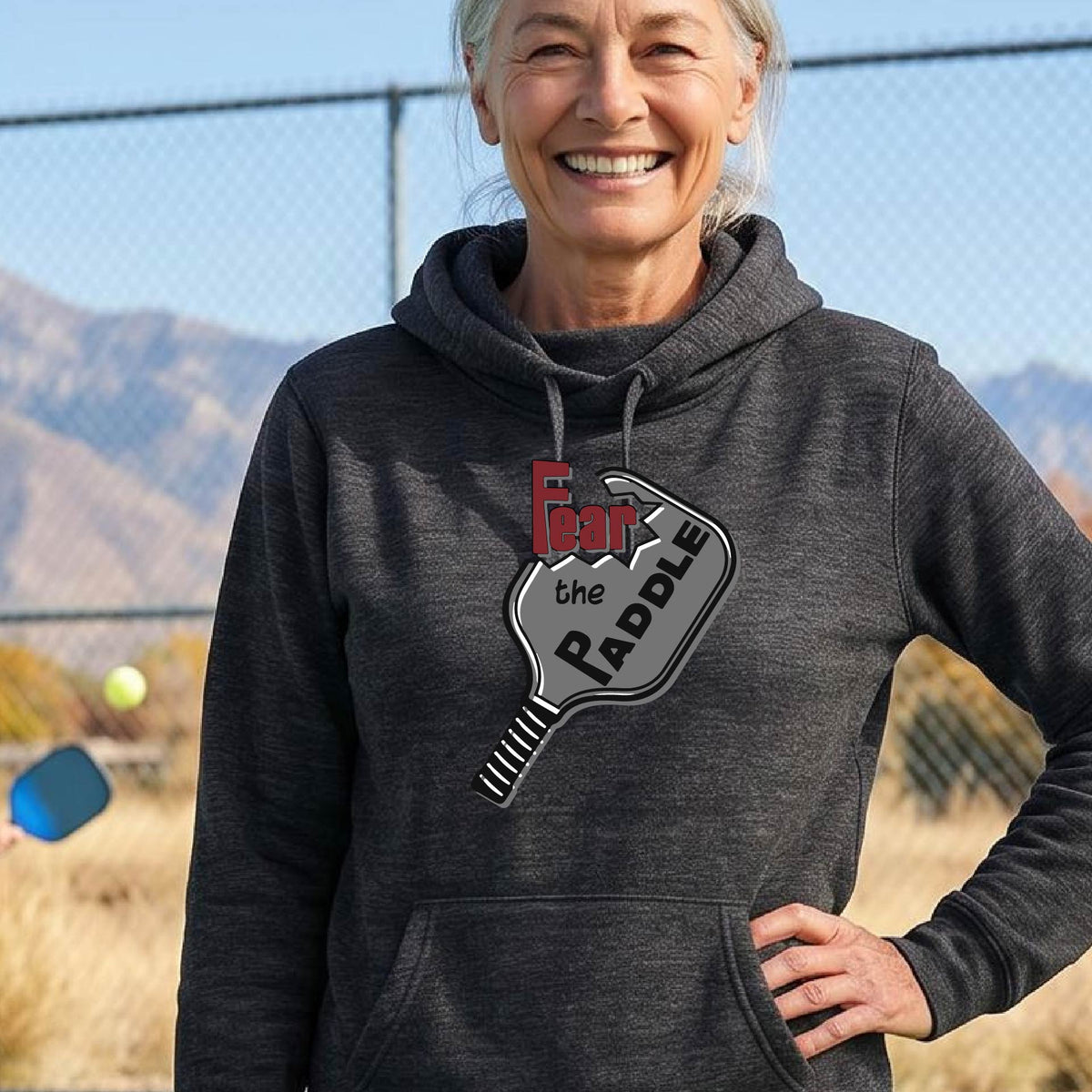 A woman wearing a black cowl-neck hoodie pullover sweatshirt with a pickleball graphic of the text 'FEAR THE PADDLE' cut into a pickleball paddle, standing on a pickleball court. 
