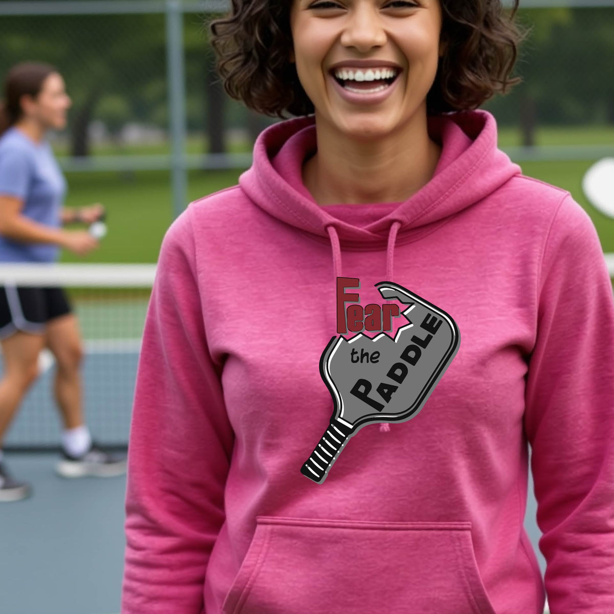 A woman wearing a pink cowl-neck hoodie pullover sweatshirt with a pickleball graphic of the text 'FEAR THE PADDLE' cut into a pickleball paddle, standing on a pickleball court. 
