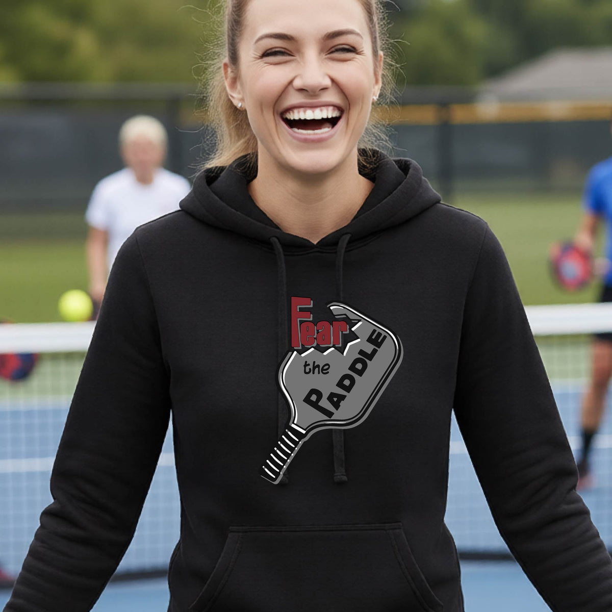 A woman wearing a black fitted hoodie featuring a pickleball graphic of the text 'FEAR THE PADDLE' cut into a pickleball paddle, standing on a pickleball court.
