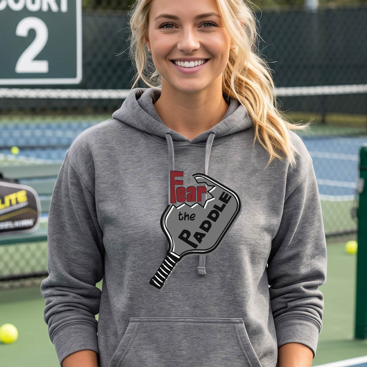 A woman wearing a gray fitted hoodie featuring a pickleball graphic of the text 'FEAR THE PADDLE' cut into a pickleball paddle, standing on a pickleball court.