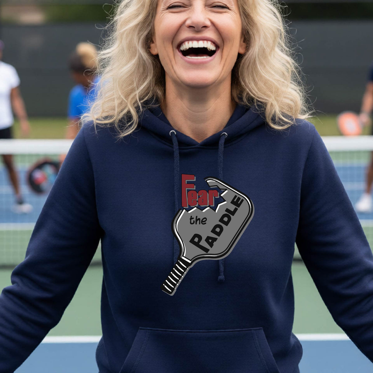 A woman wearing a navy blue fitted hoodie featuring a pickleball graphic of the text 'FEAR THE PADDLE' cut into a pickleball paddle, standing on a pickleball court.