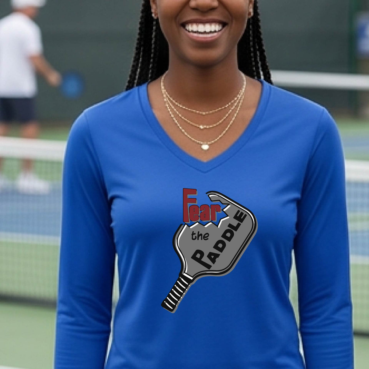 A woman on a sunny outdoor pickleball court wearing a bright royal blue long-sleeve V-neck athletic shirt with a graphic of a picklelball paddle and the text 'Fear the Paddle'.