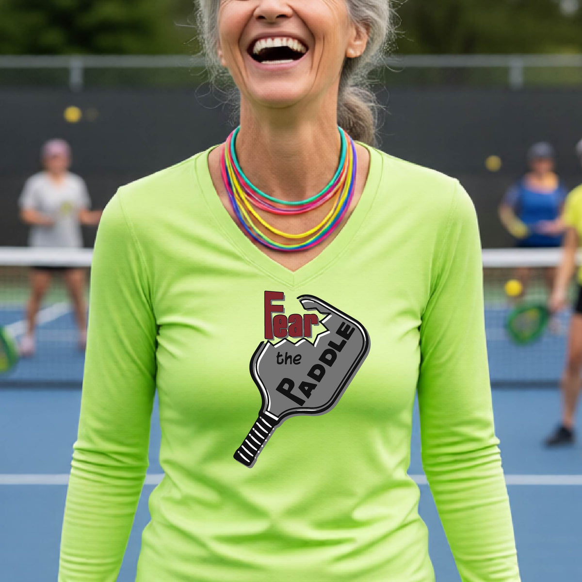 A woman on a sunny outdoor pickleball court wearing a bright lime green long-sleeve V-neck athletic shirt with a graphic of a picklelball paddle and the text 'Fear the Paddle'.