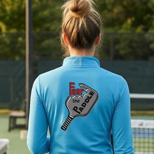 A woman wearing a bright blue quarter-zip shirt with a back pickleball graphic of the text 'FEAR THE PADDLE' cut into a pickleball paddle on the back,ona pickleball court. 