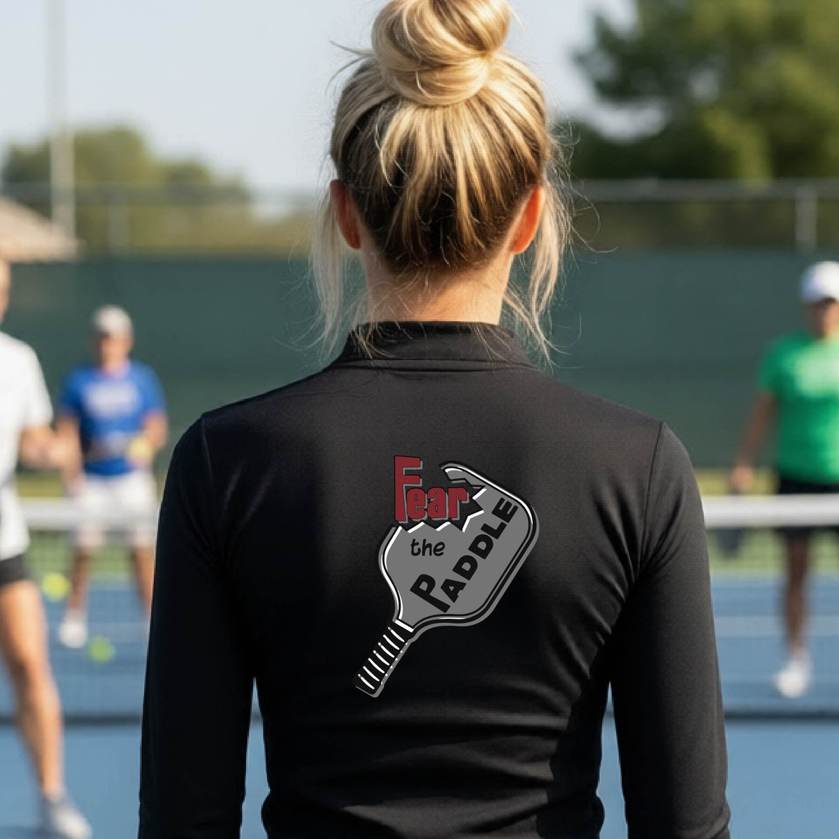 A woman wearing a black quarter-zip shirt with a back pickleball graphic of the text 'FEAR THE PADDLE' cut into a pickleball paddle on the back,ona pickleball court. 