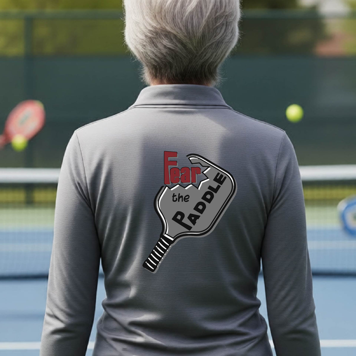 A woman wearing a gray quarter-zip shirt with a back pickleball graphic of the text 'FEAR THE PADDLE' cut into a pickleball paddle on the back,ona pickleball court. 
