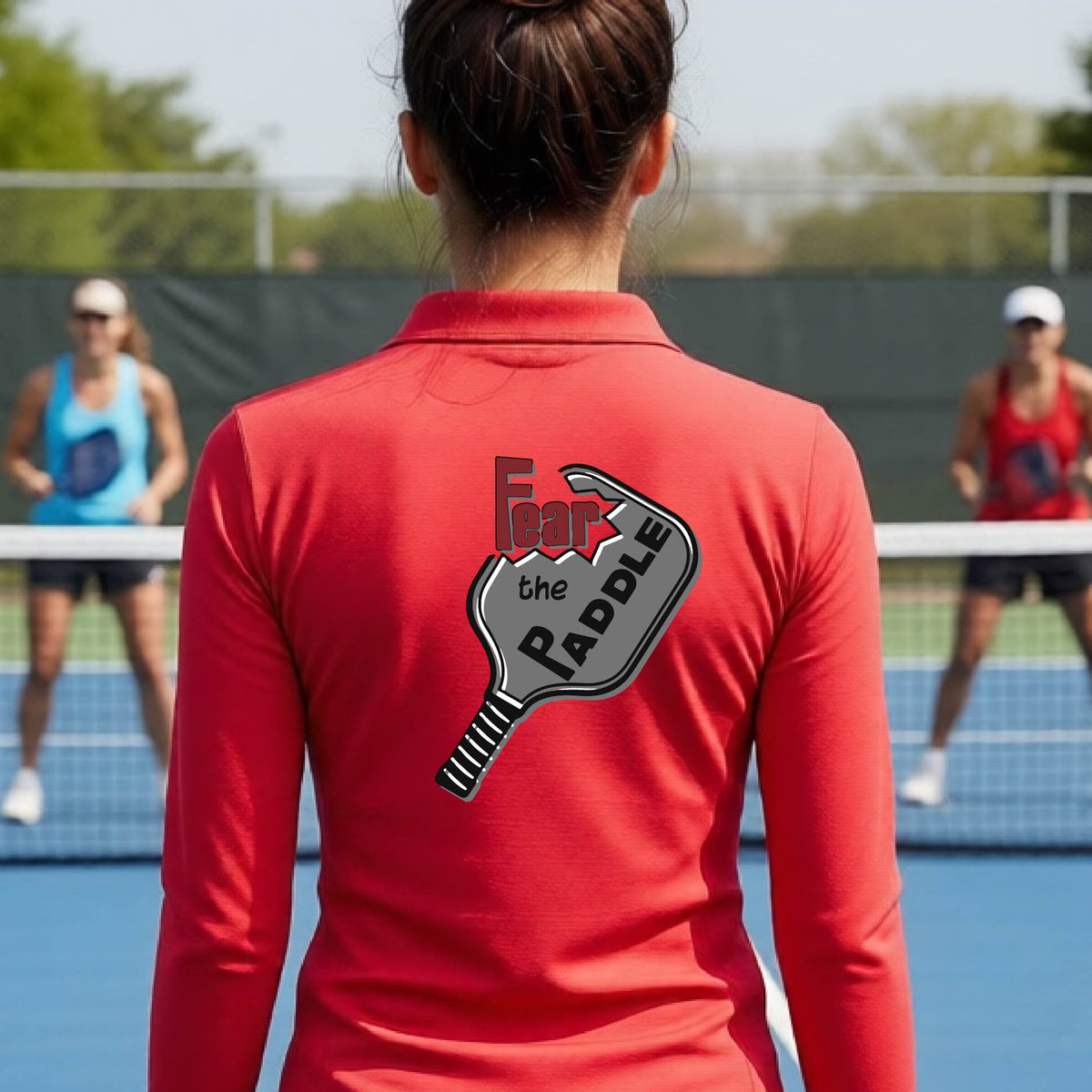 A woman wearing a red quarter-zip shirt with a back pickleball graphic of the text 'FEAR THE PADDLE' cut into a pickleball paddle on the back,ona pickleball court. 