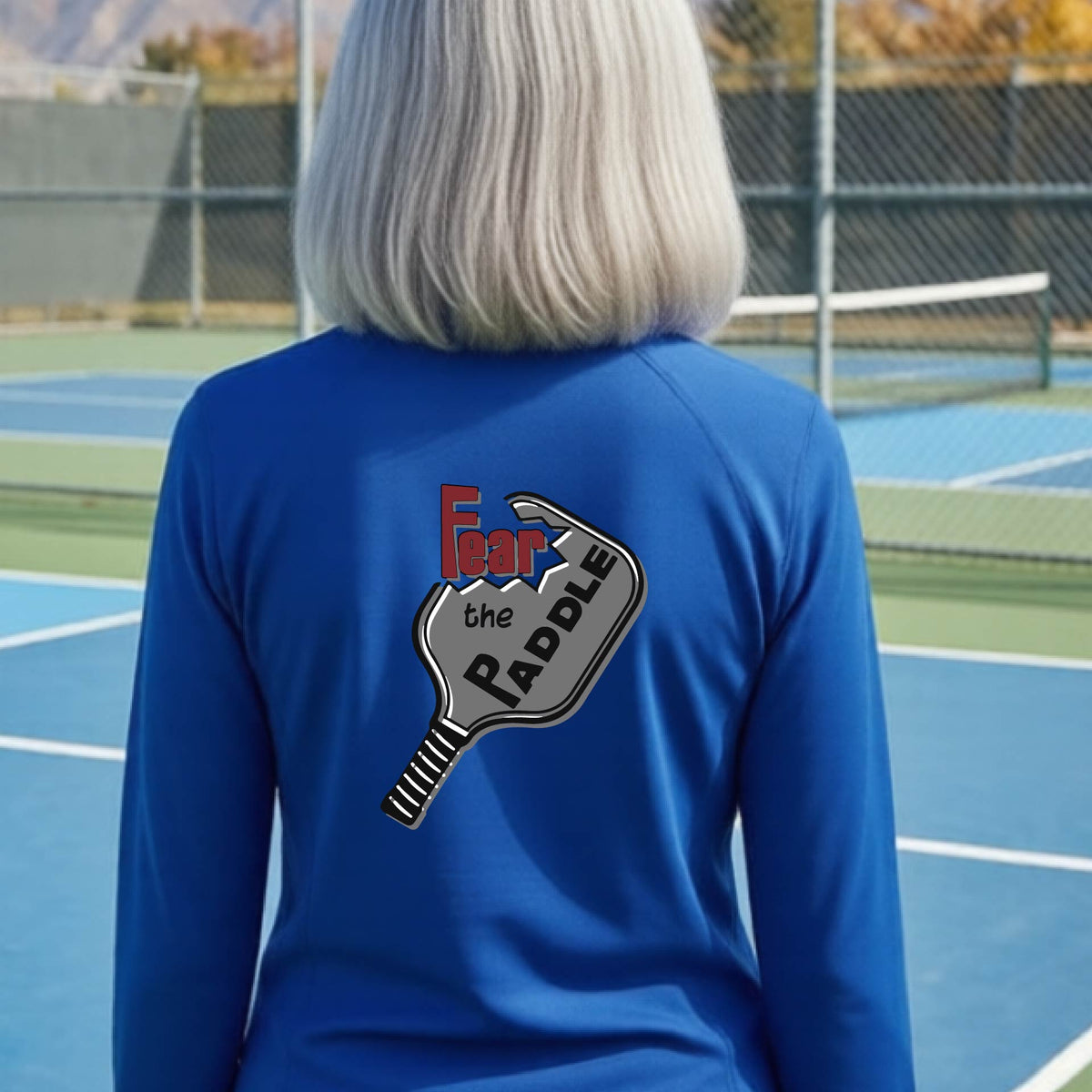 A woman wearing a bright royal blue quarter-zip shirt with a back pickleball graphic of the text 'FEAR THE PADDLE' cut into a pickleball paddle on the back,ona pickleball court. 