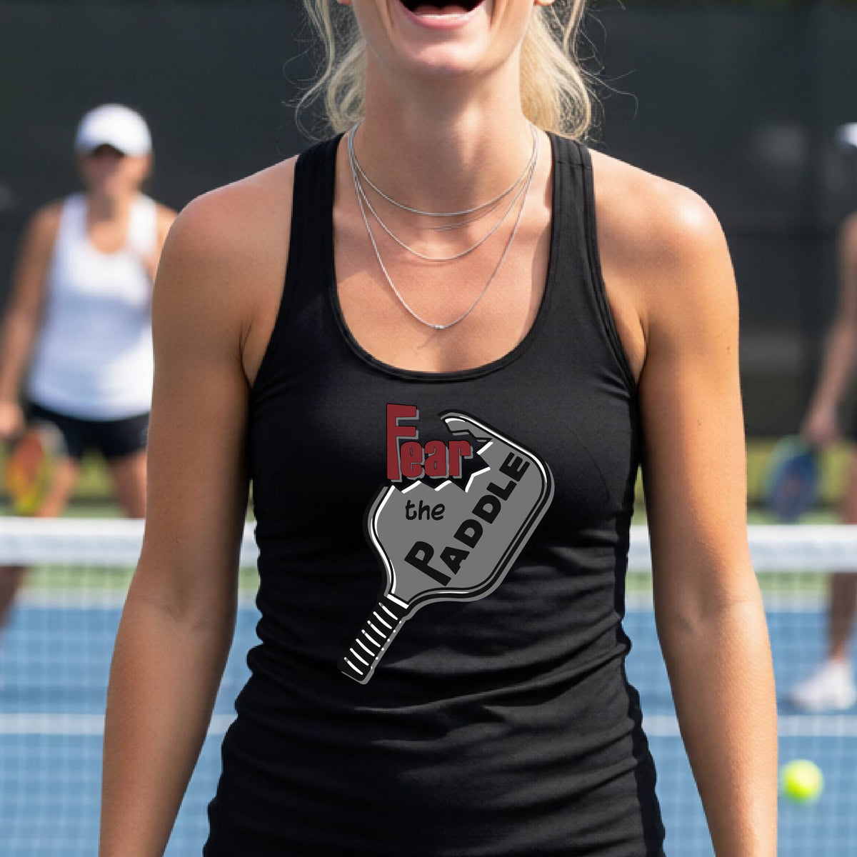 A woman on a pickleball court wearing a black athletic tank top with a graphic that reads 'Fear the Paddle' over a pickleball paddle illustration.