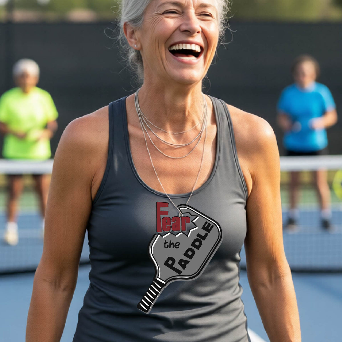 A woman on a pickleball court wearing a gray athletic tank top with a graphic that reads 'Fear the Paddle' over a pickleball paddle illustration.