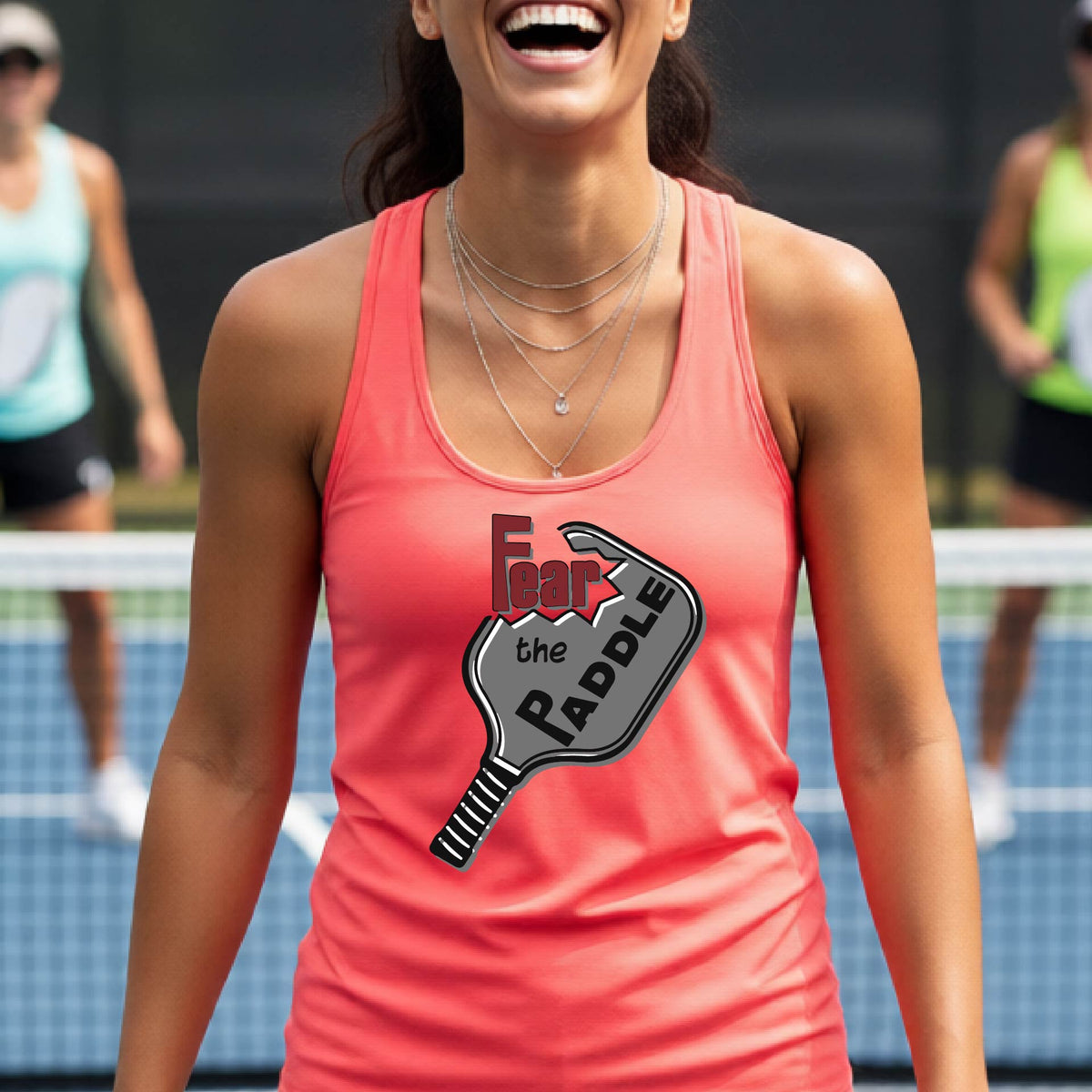 A woman on a pickleball court wearing a coral pink athletic tank top with a graphic that reads 'Fear the Paddle' over a pickleball paddle illustration.