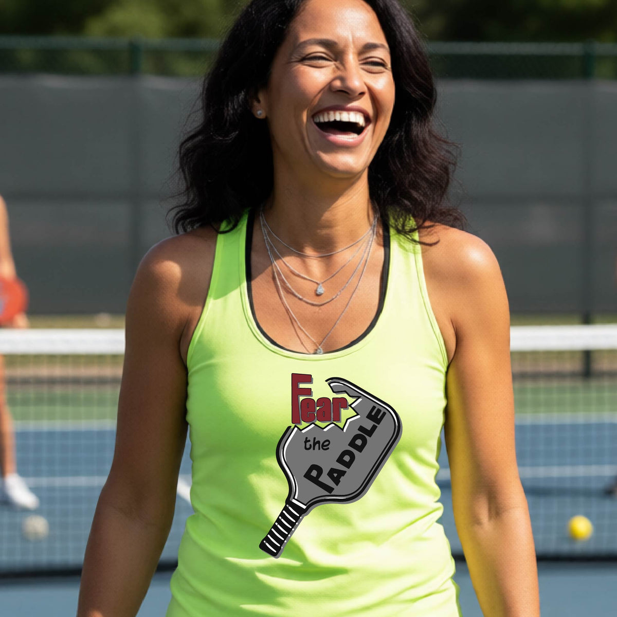 A woman on a pickleball court wearing a lime green athletic tank top with a graphic that reads 'Fear the Paddle' over a pickleball paddle illustration.
