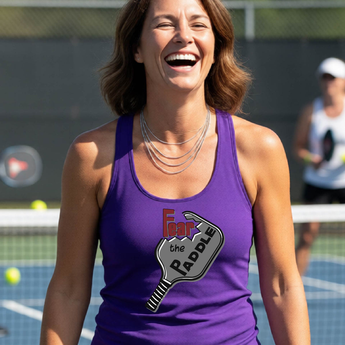 A woman on a pickleball court wearing a purple athletic tank top with a graphic that reads 'Fear the Paddle' over a pickleball paddle illustration.