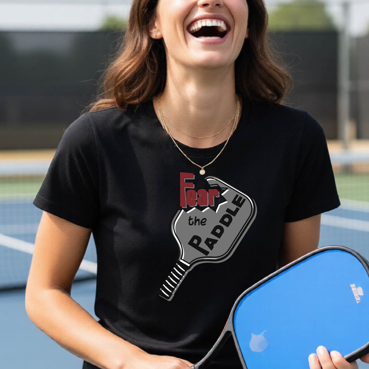A woman laughing on a pickleball court wearing a black t-shirt with a graphic that reads 'Fear the PADDLE' over a paddle-shaped design. 