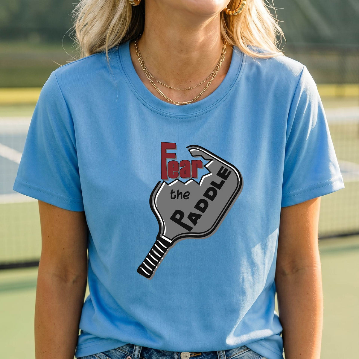 A woman laughing on a pickleball court wearing a blue t-shirt with a graphic that reads 'Fear the PADDLE' over a paddle-shaped design. 