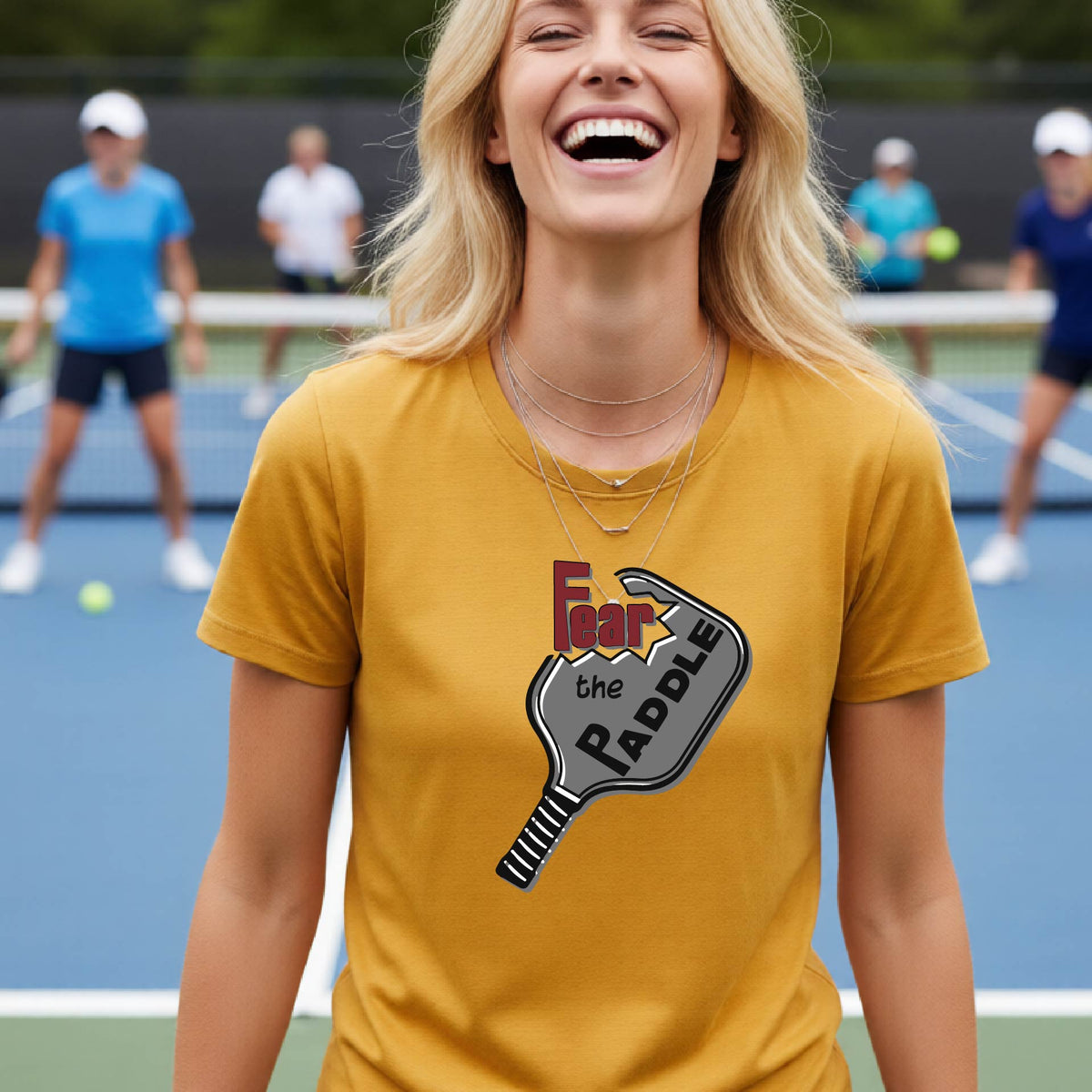 A woman laughing on a pickleball court wearing a mustard yellow gold t-shirt with a graphic that reads 'Fear the PADDLE' over a paddle-shaped design. 