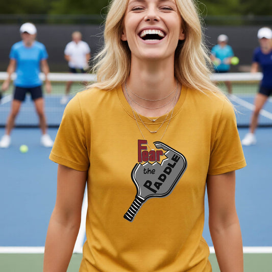 A woman laughing on a pickleball court wearing a mustard yellow gold t-shirt with a graphic that reads 'Fear the PADDLE' over a paddle-shaped design. 
