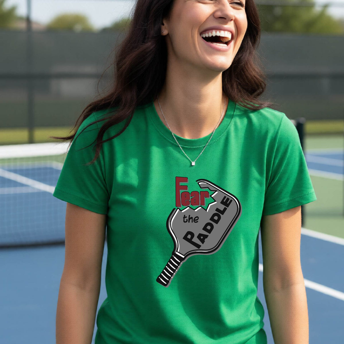 A woman laughing on a pickleball court wearing a green t-shirt with a graphic that reads 'Fear the PADDLE' over a paddle-shaped design. 