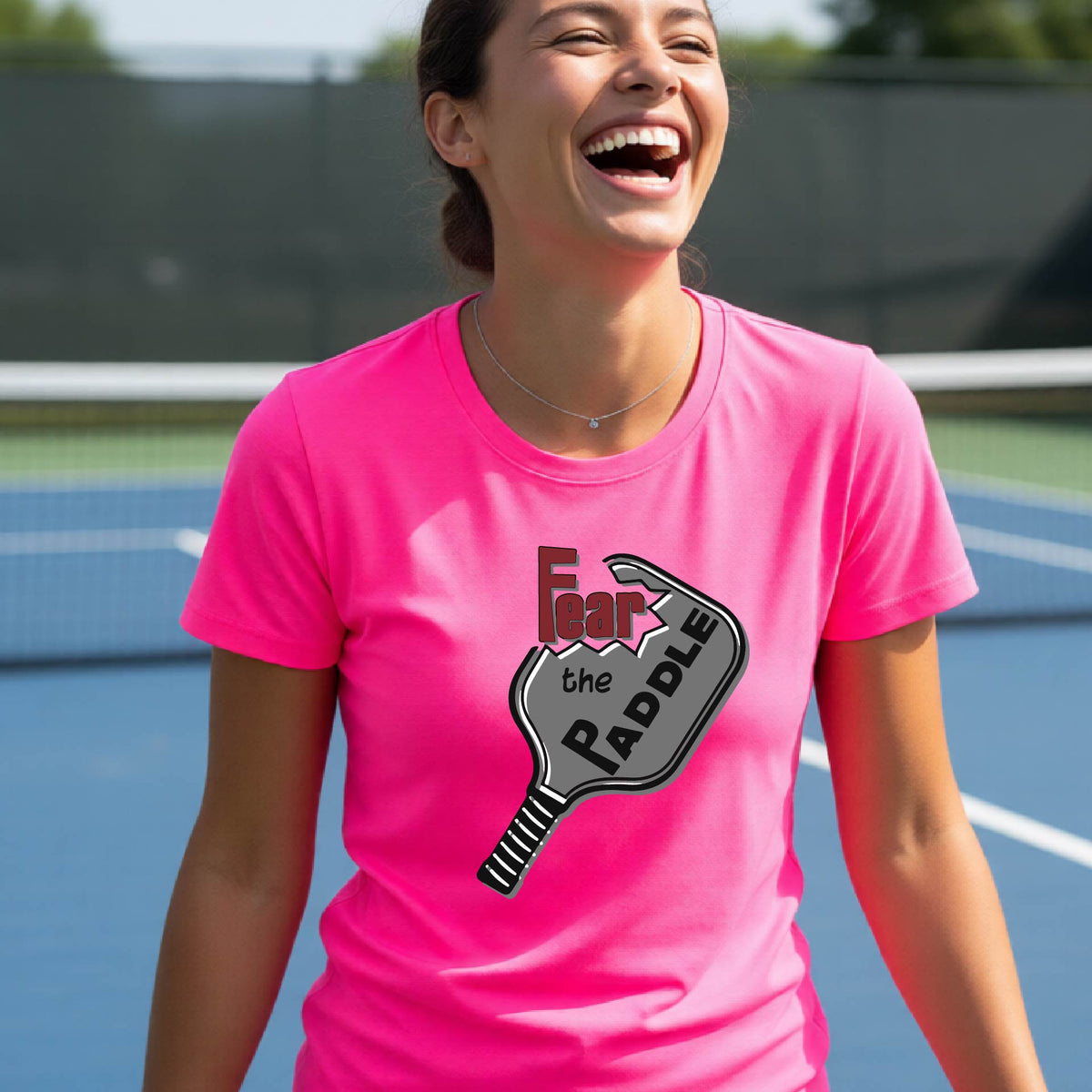 A woman laughing on a pickleball court wearing a pink t-shirt with a graphic that reads 'Fear the PADDLE' over a paddle-shaped design. 