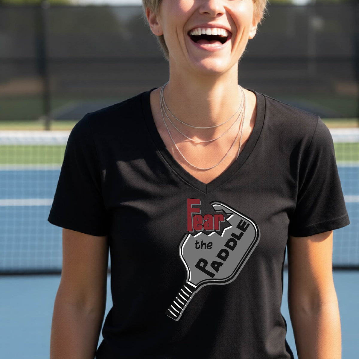 A woman on a pickleball court wearing a black V-neck t-shirt with a graphic that reads 'Fear the Paddle' in red and black over a gray pickleball paddle illustration.