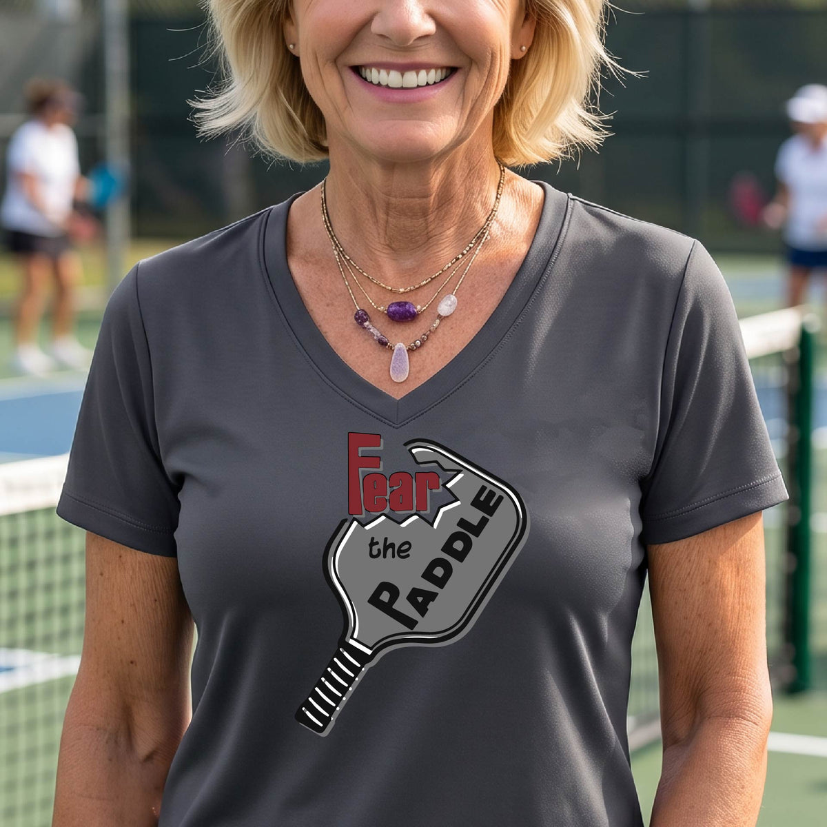 A woman on a pickleball court wearing a gray V-neck t-shirt with a graphic that reads 'Fear the Paddle' in red and black over a gray pickleball paddle illustration.