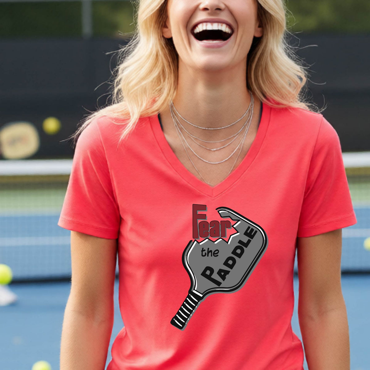 A woman on a pickleball court wearing a coral pink V-neck t-shirt with a graphic that reads 'Fear the Paddle' in red and black over a gray pickleball paddle illustration.