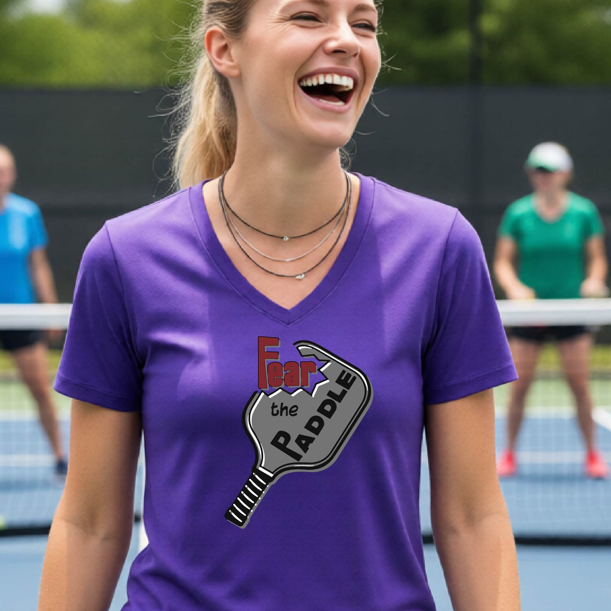 A woman on a pickleball court wearing a purple V-neck t-shirt with a graphic that reads 'Fear the Paddle' in red and black over a gray pickleball paddle illustration.
