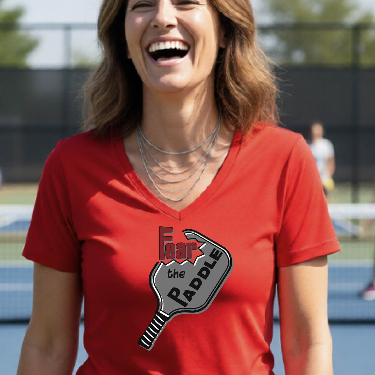 A woman on a pickleball court wearing a red V-neck t-shirt with a graphic that reads 'Fear the Paddle' in red and black over a gray pickleball paddle illustration.