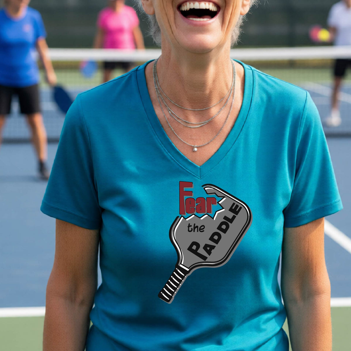 A woman on a pickleball court wearing a teal V-neck t-shirt with a graphic that reads 'Fear the Paddle' in red and black over a gray pickleball paddle illustration.
