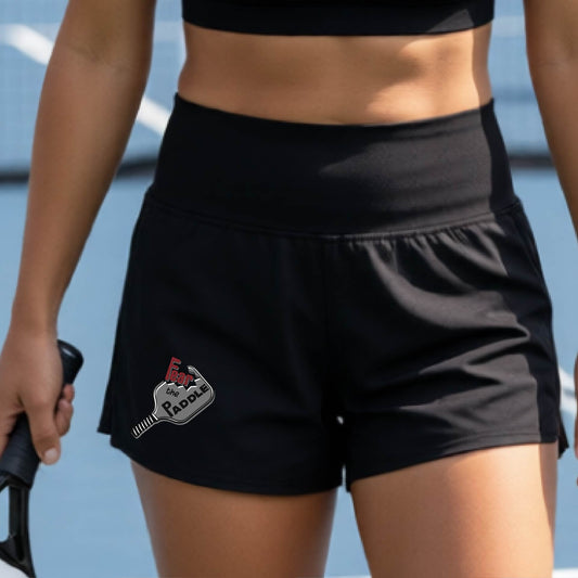 A woman wearing black athletic shorts featuring a pickleball graphic of the text 'FEAR THE PADDLE' cut into a pickleball paddle onm the lower left of the shorts, standing on a pickleball court.
