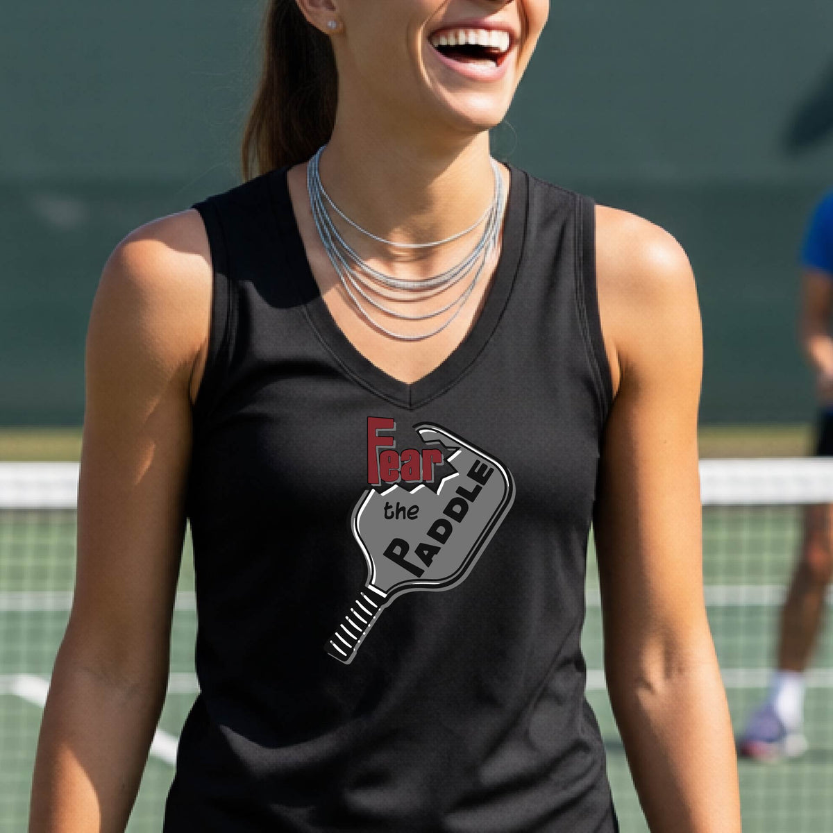 A woman wearing a black sleeveless tank top featuring a pickleball graphic of the text 'FEAR THE PADDLE' cut into a pickleball paddle, standing on a pickleball court. 