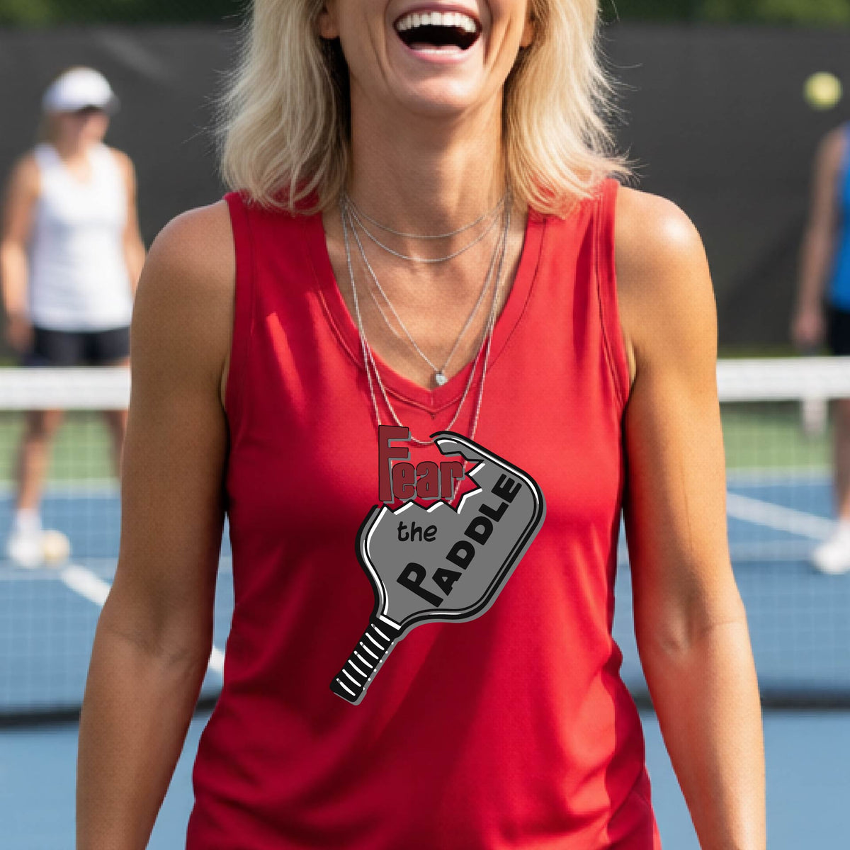 A woman wearing a red sleeveless tank top featuring a pickleball graphic of the text 'FEAR THE PADDLE' cut into a pickleball paddle, standing on a pickleball court. 