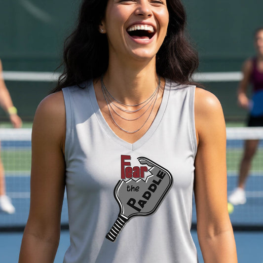 A woman wearing a silver gray sleeveless tank top featuring a pickleball graphic of the text 'FEAR THE PADDLE' cut into a pickleball paddle, standing on a pickleball court. 