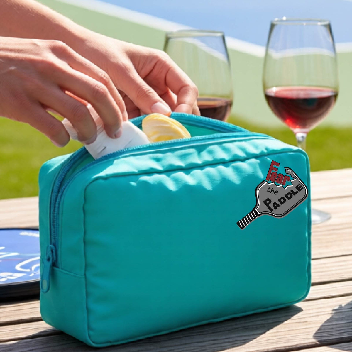 An aqua turquoise cosmetic bag with a zippered closure and a pickleball graphic of the text 'FEAR THE PADDLE' with the words cut into a pickleball paddle. 