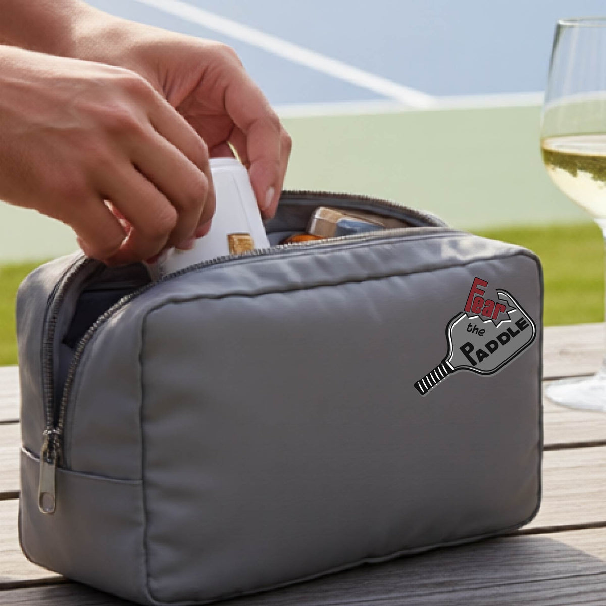 A gray cosmetic bag with a zippered closure and a pickleball graphic of the text 'FEAR THE PADDLE' with the words cut into a pickleball paddle. 