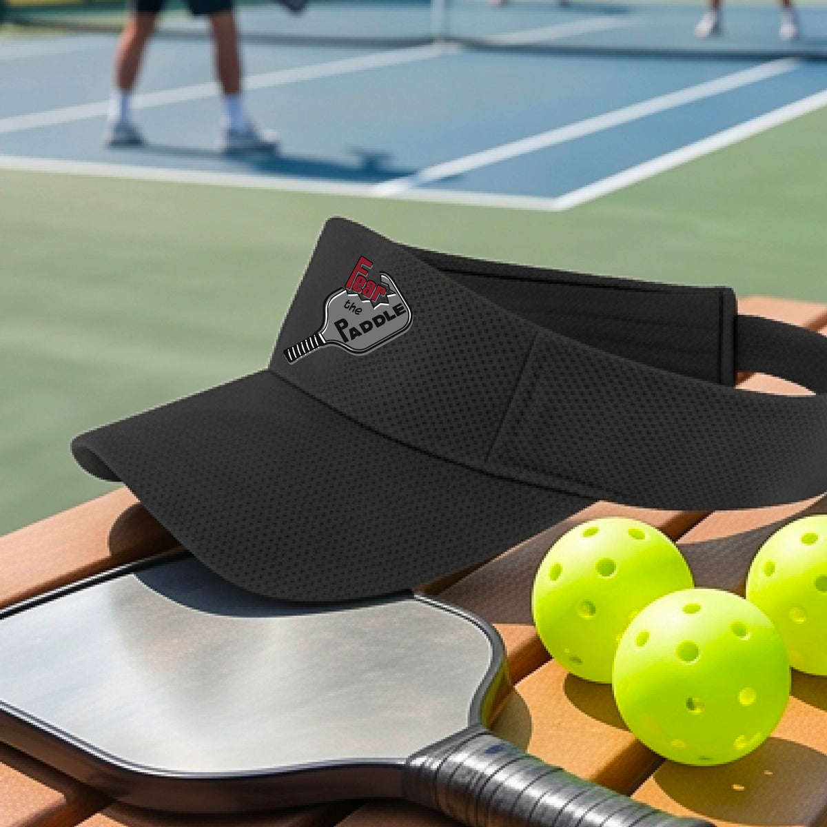 A close up of a black visor with a 'Fear the Paddle' cutout from a pickleall paddle with a blurred pickleblal court in the background.