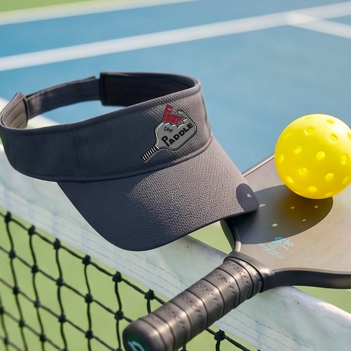 A close up of a graphite gray visor with a 'Fear the Paddle' cutout from a pickleall paddle with a blurred pickleblal court in the background.