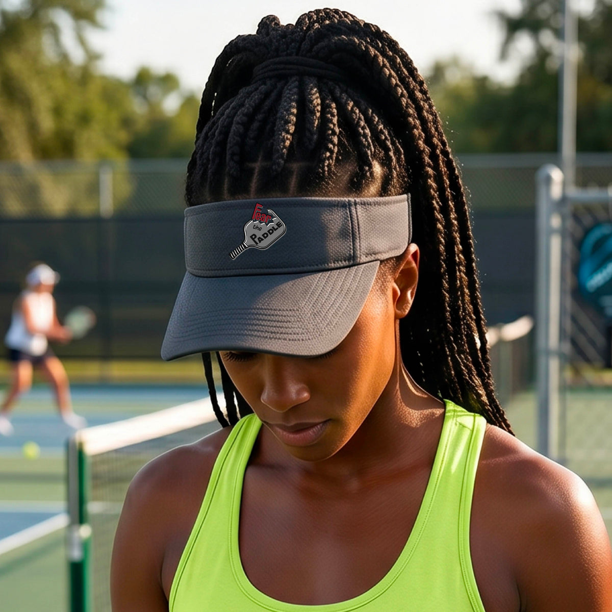 A close up of a graphite visor with a 'Fear the Paddle' cutout from a pickleall paddle with a blurred pickleblal court in the background.