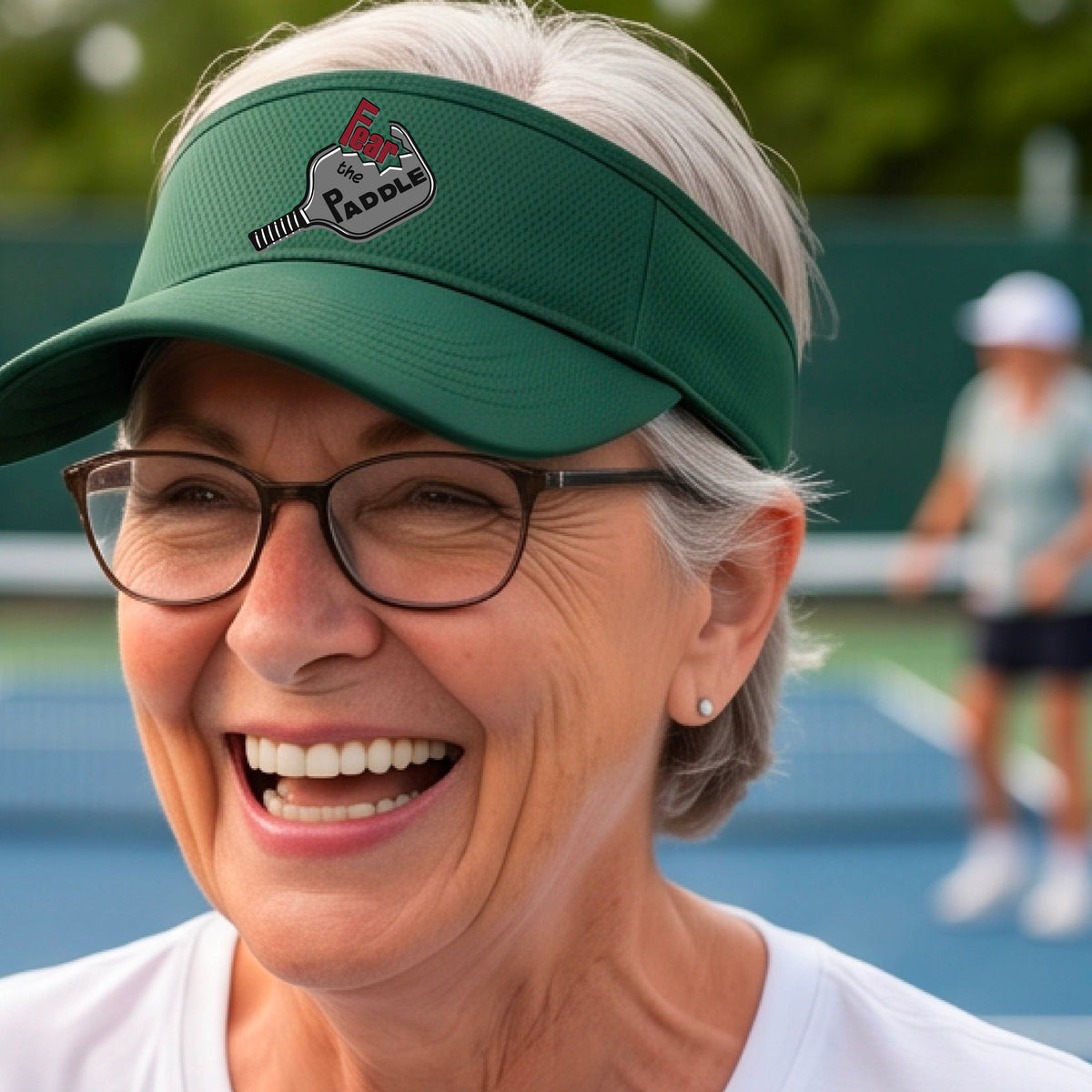 A close up of a green visor with a 'Fear the Paddle' cutout from a pickleall paddle with a blurred pickleblal court in the background.