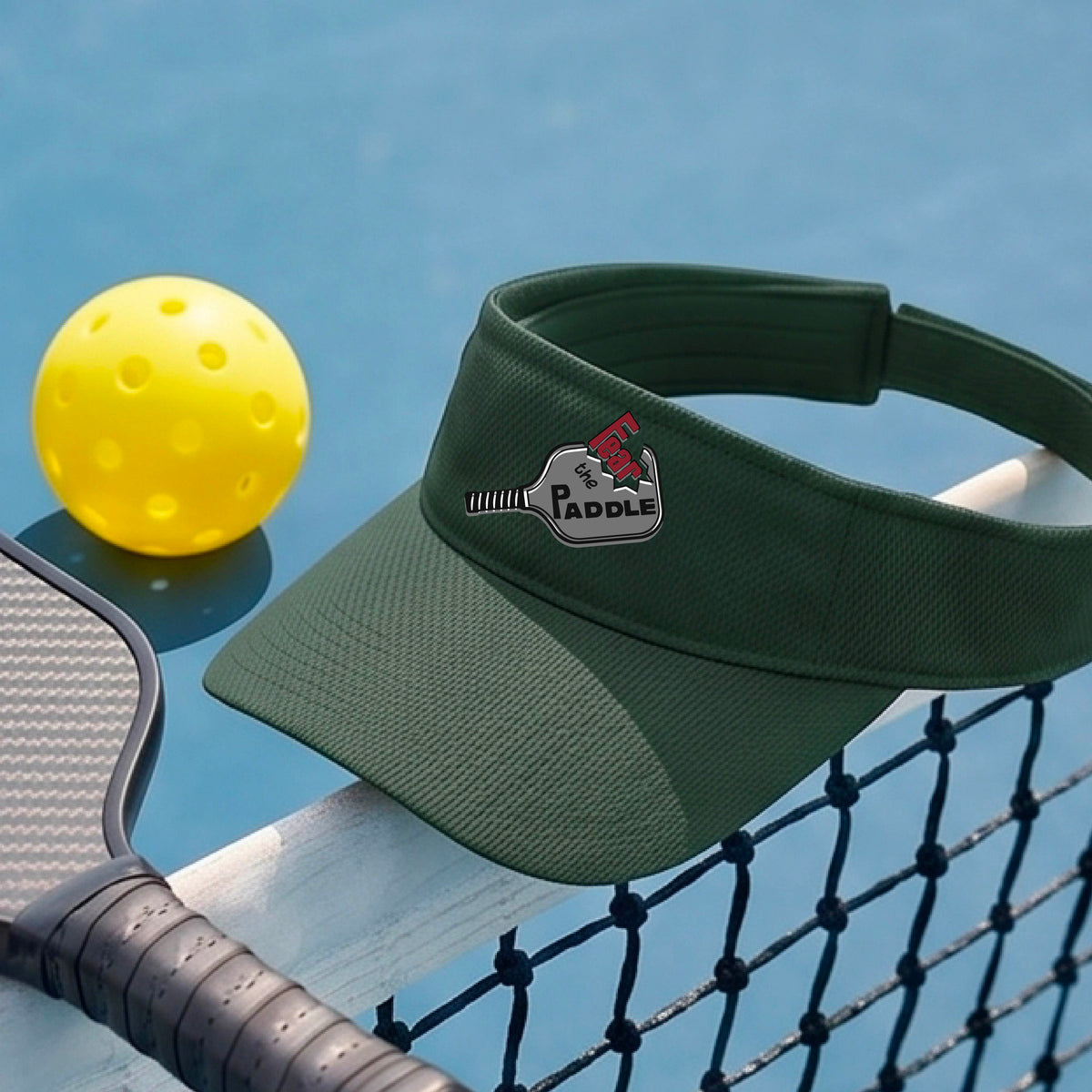 A close up of a green visor with a 'Fear the Paddle' cutout from a pickleall paddle with a blurred pickleblal court in the background.