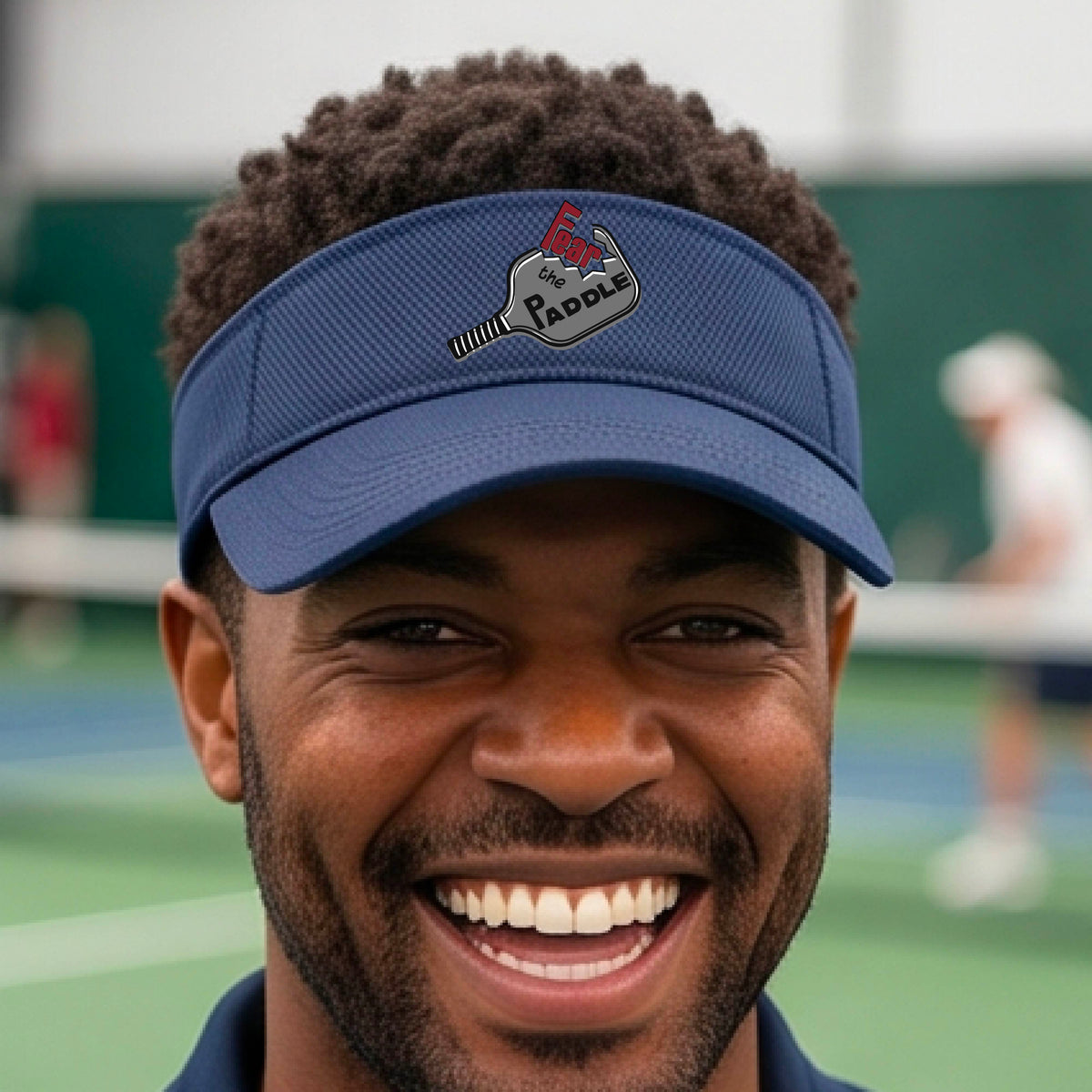 A close up of a navy blue visor with a 'Fear the Paddle' cutout from a pickleall paddle with a blurred pickleblal court in the background.