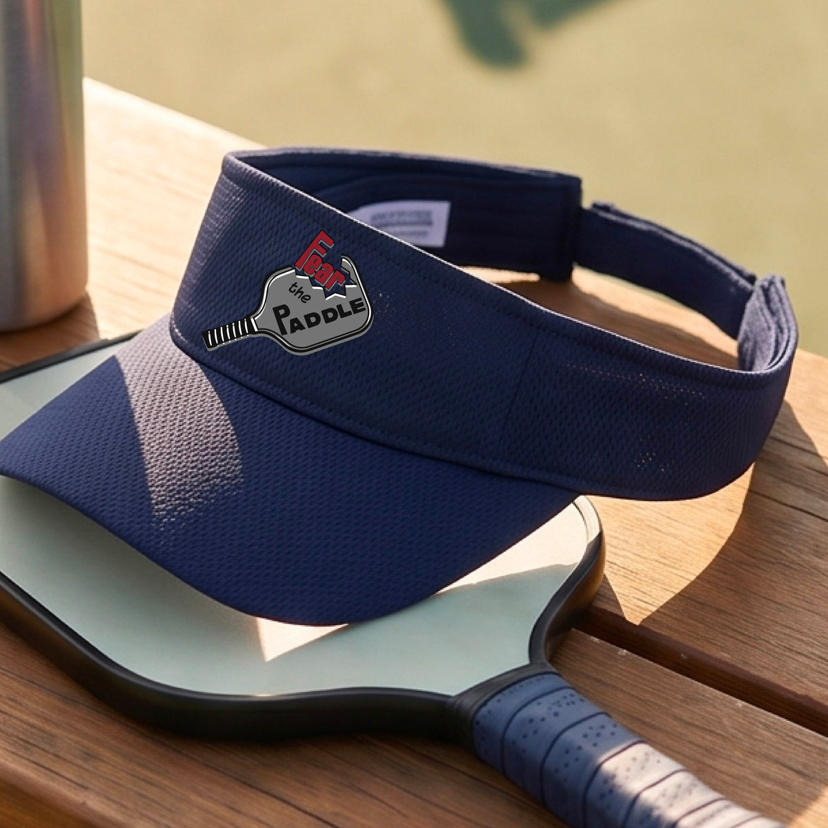 A close up of a navy blue visor with a 'Fear the Paddle' cutout from a pickleall paddle with a blurred pickleblal court in the background.