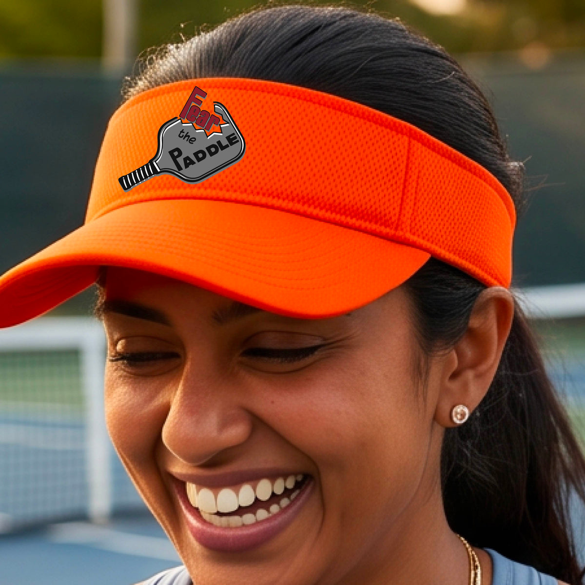 A close up of a bright orange visor with a 'Fear the Paddle' cutout from a pickleall paddle with a blurred pickleblal court in the background.