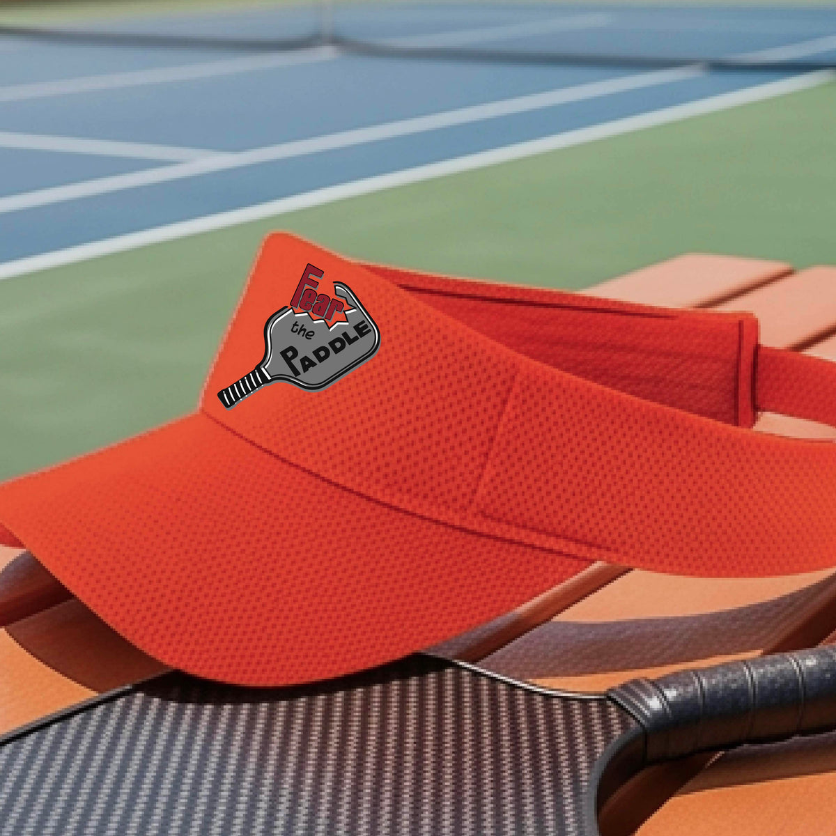 A close up of an orange visor with a 'Fear the Paddle' cutout from a pickleall paddle with a blurred pickleblal court in the background.