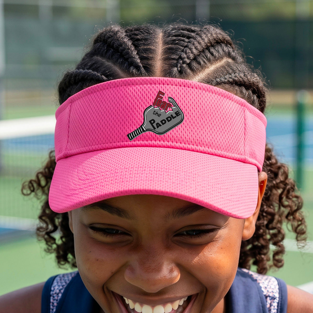 A close up of a pink visor with a 'Fear the Paddle' cutout from a pickleall paddle with a blurred pickleblal court in the background.
