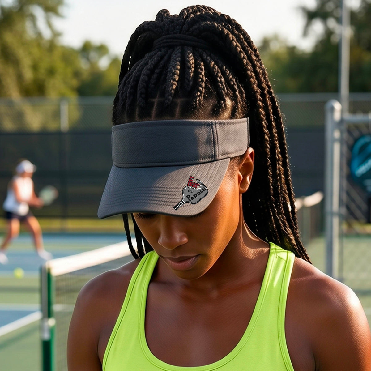 A close up of a graphite visor with a 'Fear the Paddle' cutout from a pickleall paddle with a blurred pickleblal court in the background.